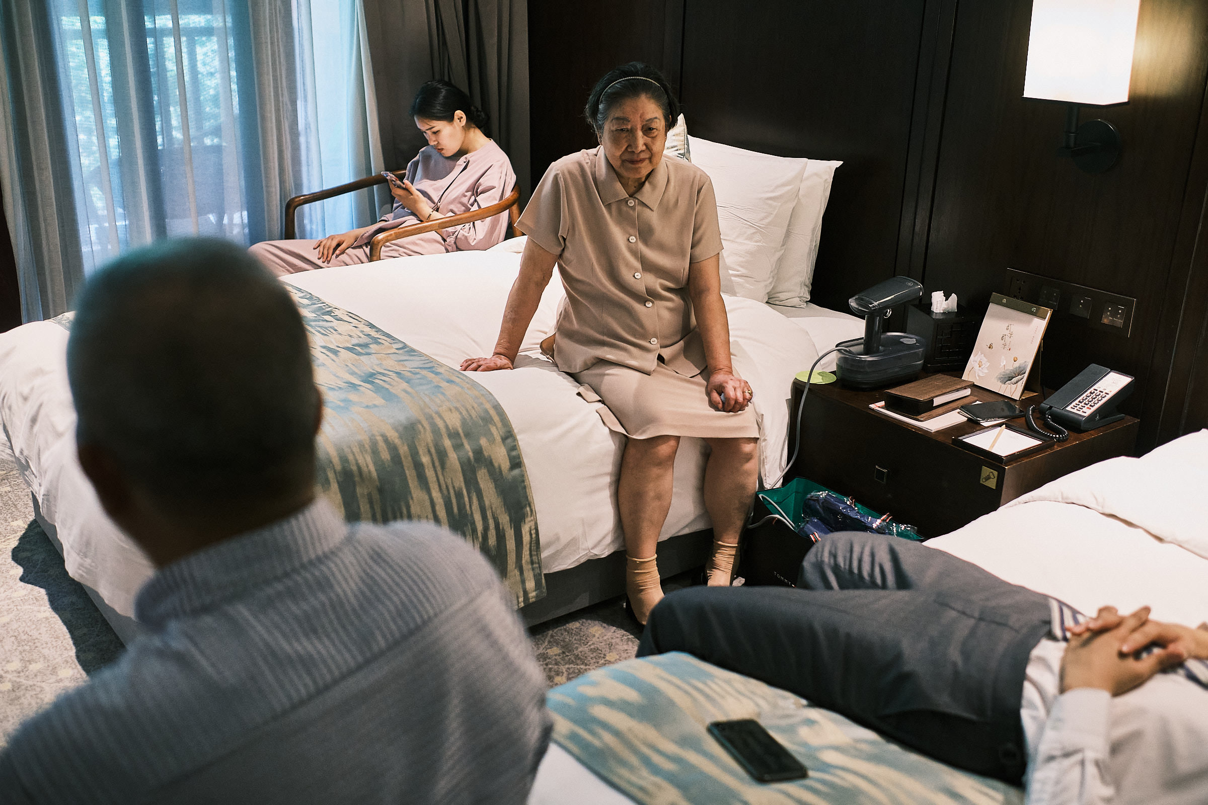 Grandmother Sitting On Bed While Figure Of A Man Lays On Bed And Father Of The Bride Holds A Conversation The Morning Of The Wedding In Hangzhou China