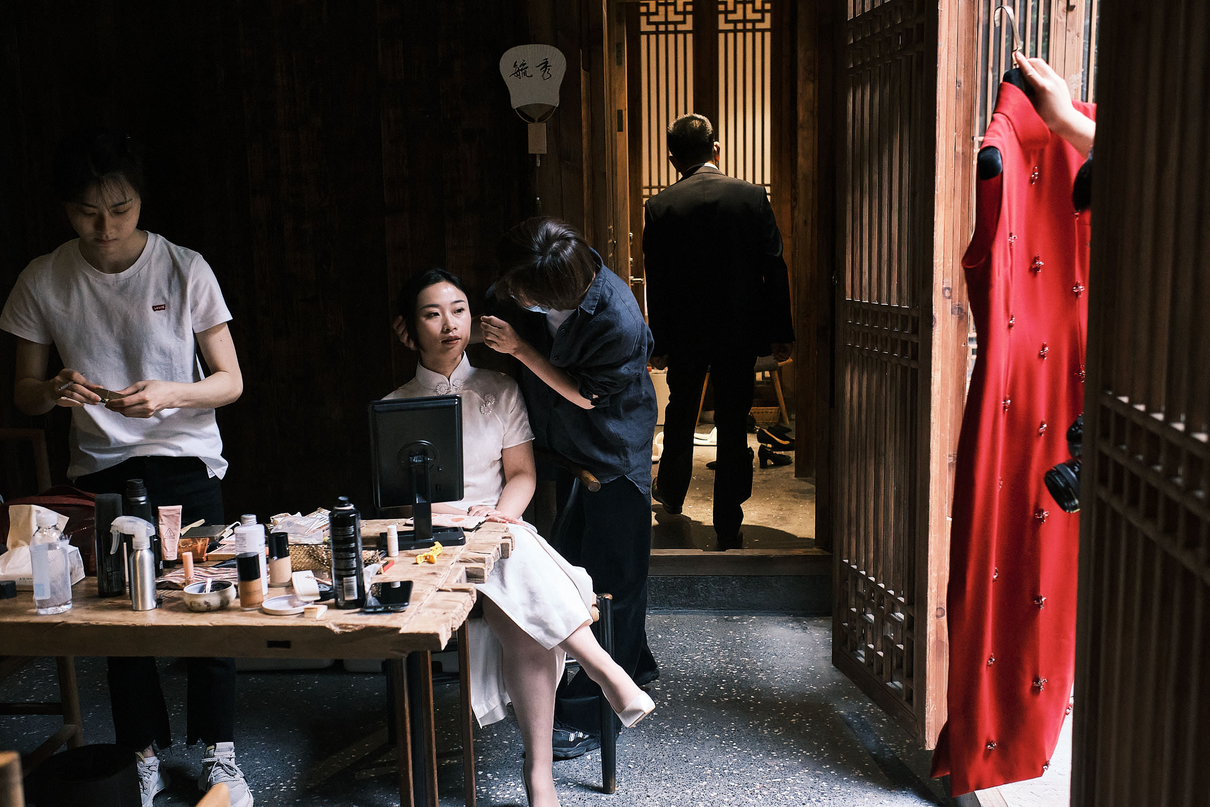 Bride Does Her Makeup As Someone Holds Red Dress Through Door Frame And Father Walks Away In Hangzhou China