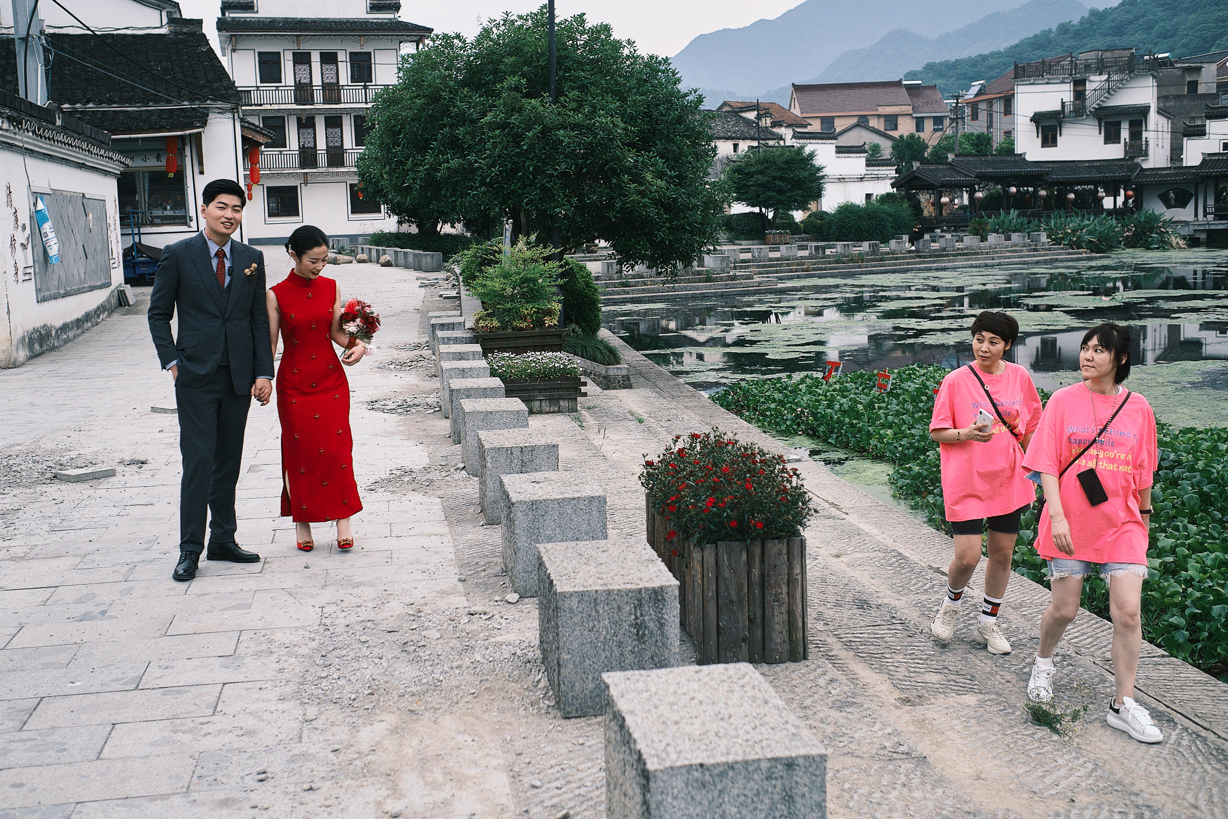 Bride Dressed In Red And Groom Stand Next To The Lake As Tourists Dressed In Pink Pass By