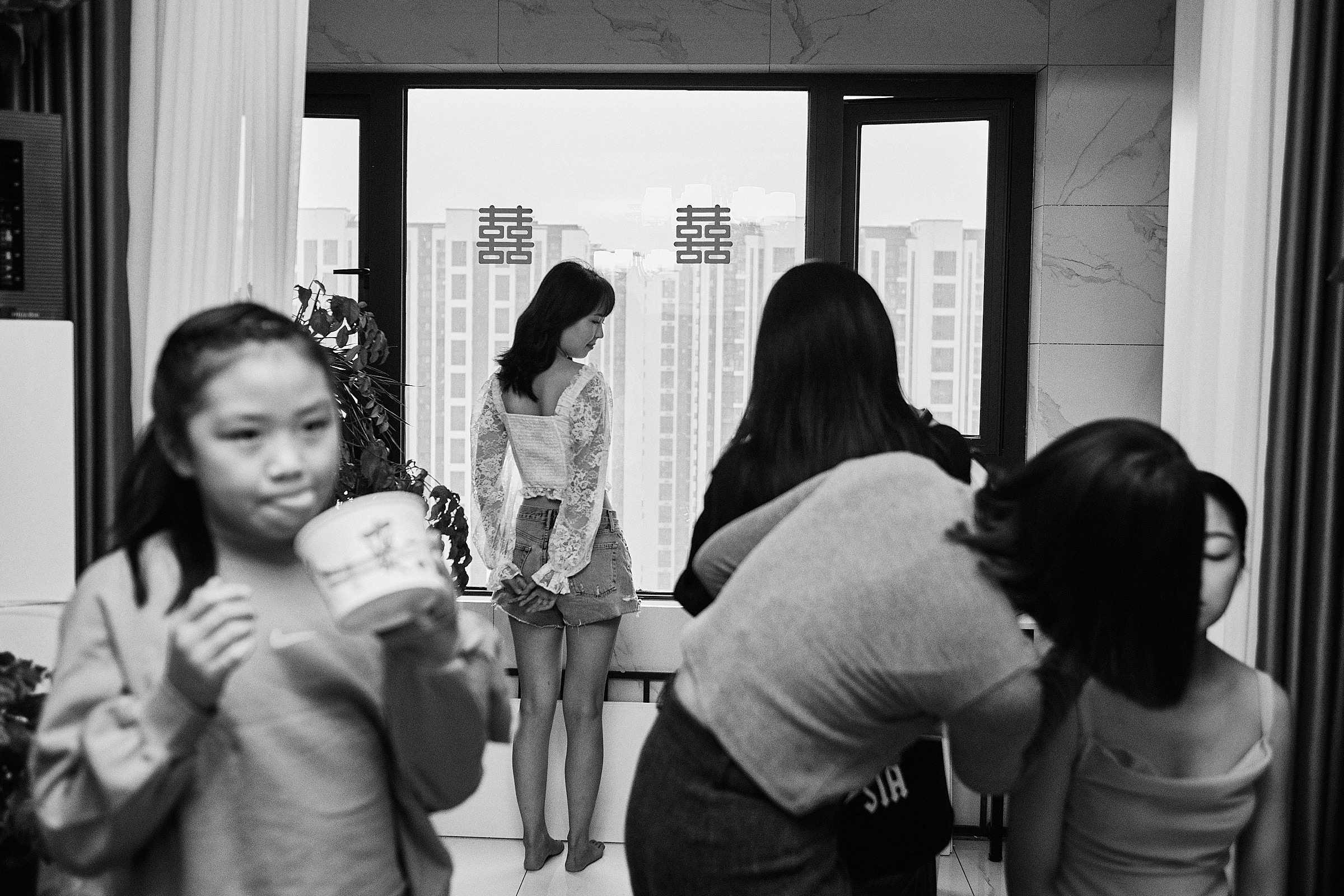 Bride Stands By The Window As Her Sister Eats Instant Noodles In Black And White