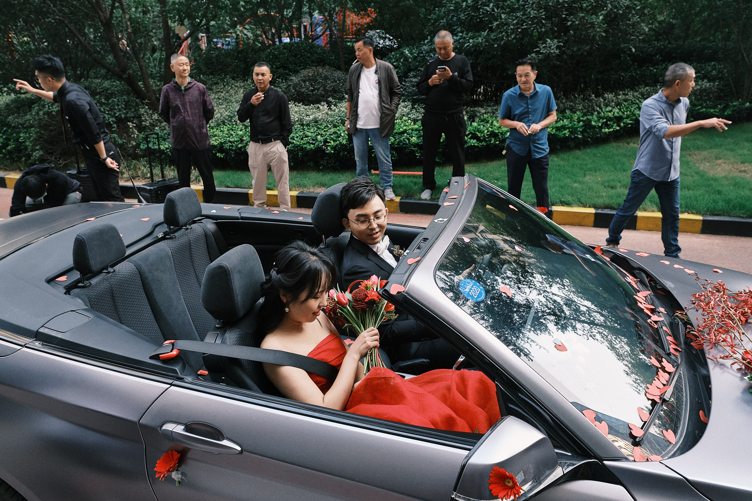 Bride Dressed In Red And Groom Await In The Car As Family Members Stand Behind In Symmetry