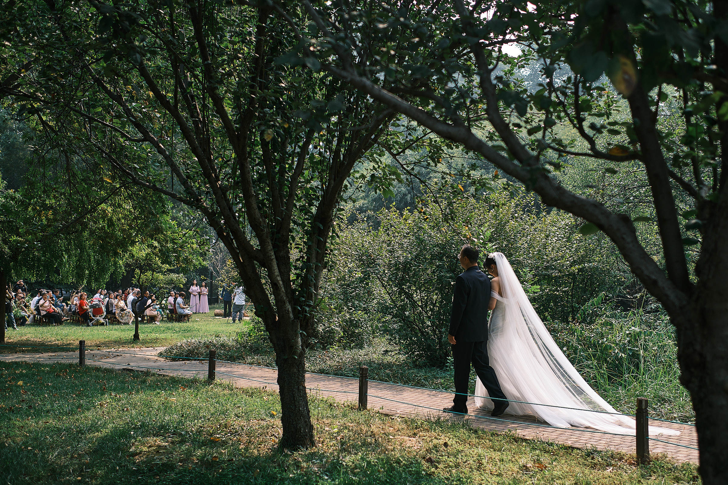 Bride And Her Father Walk Towards The Ceremony Location In Exterior Framed By Trees And Green Scenery