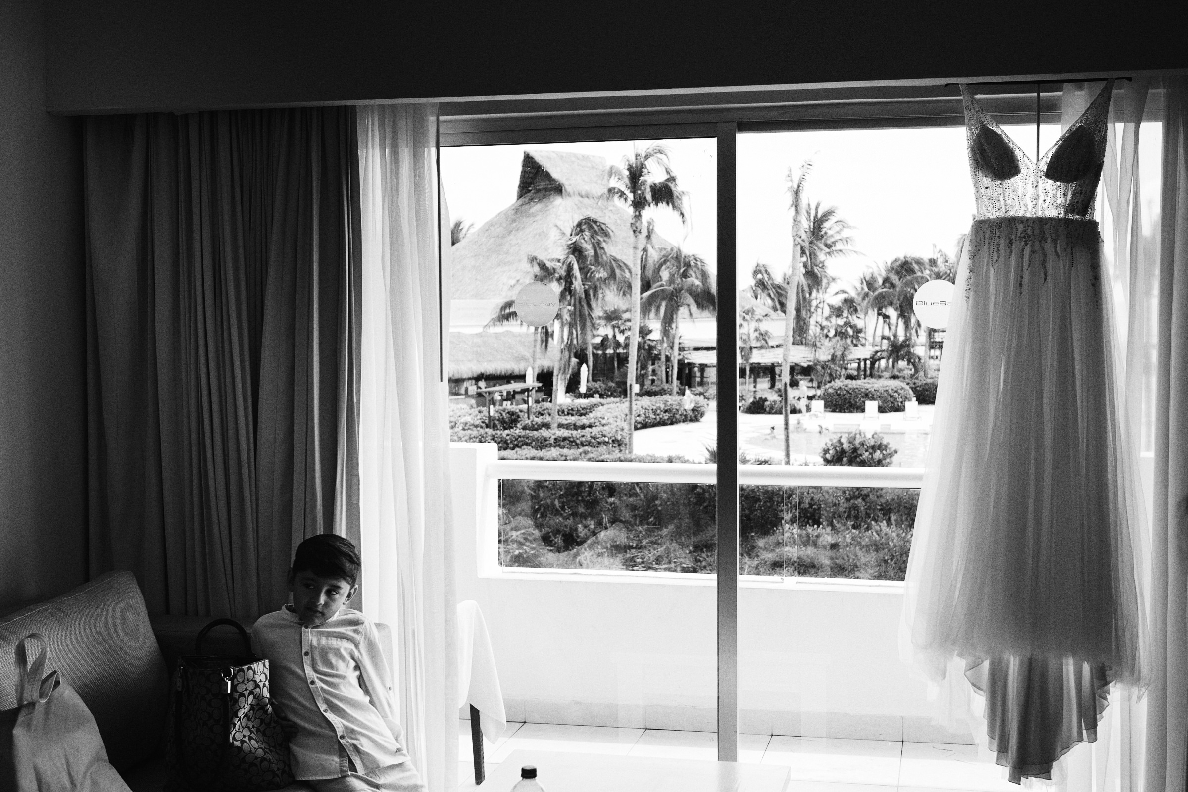 Son Of Bride Looks Into Room As Bridal Dress Hangs By The Window