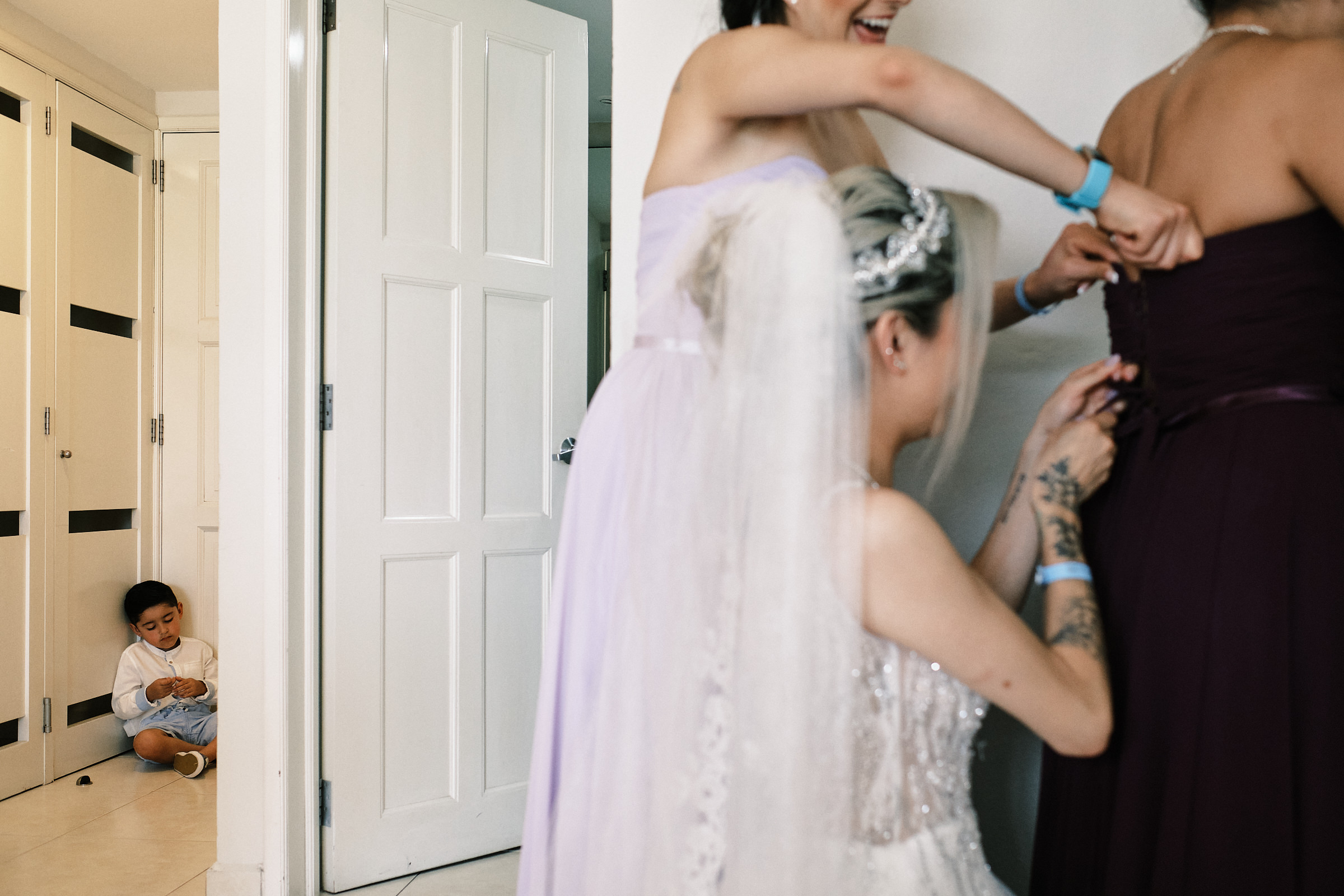 Son Of The Bride Awaits Next To Door As Mom Helps Bridesmaids Get Ready