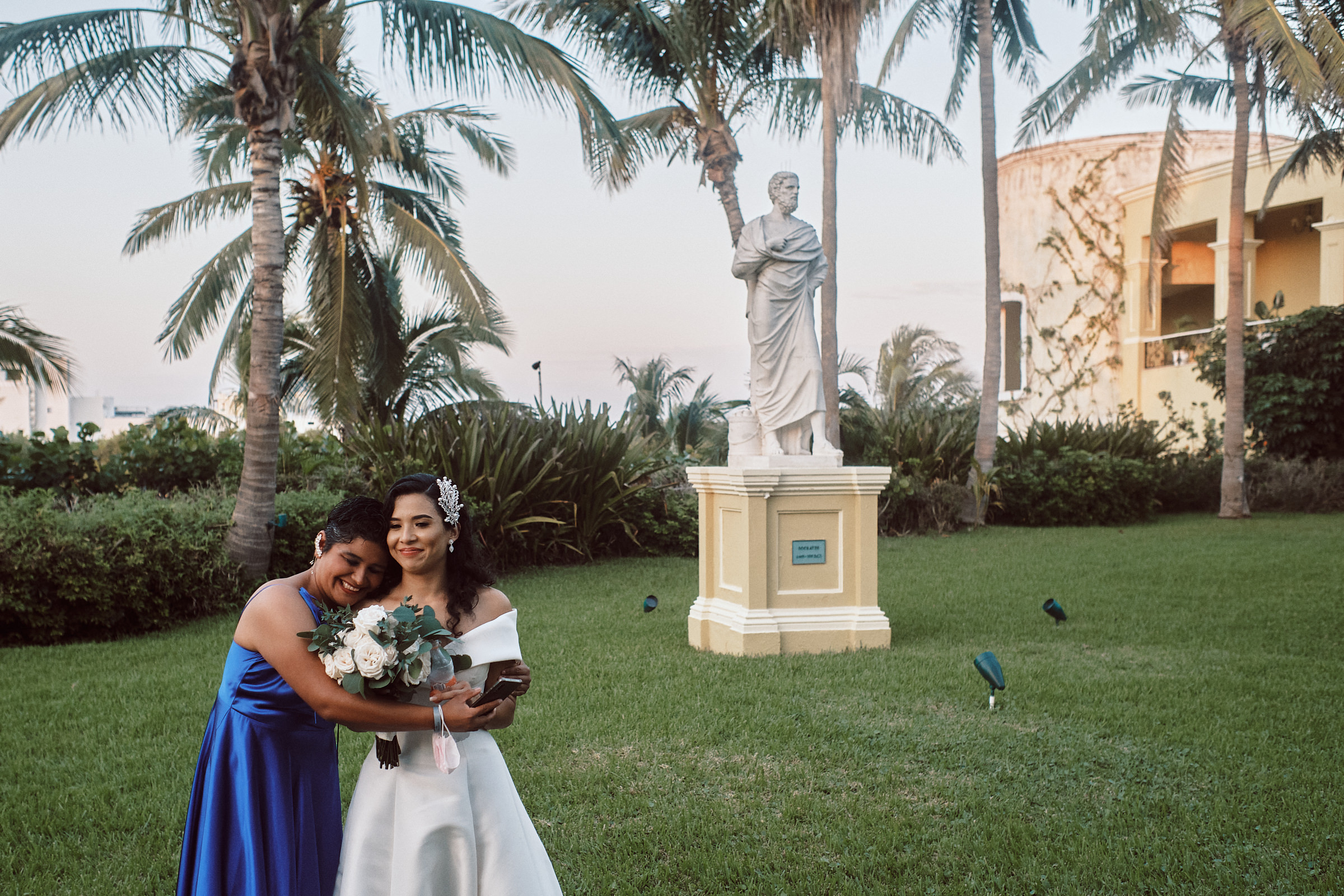 Bride Receives Hug By Her Friend In Front Of Statue At Hotel Pueblo Bonito Emerald Bay In Mexico